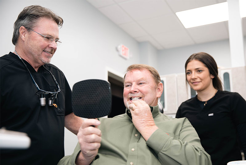 Dr. Chuck Pitts And Refresh Me Dental Center Staff Showing A Dental Implant Patient His New Mouth With A Mirror After The Implant Surgery